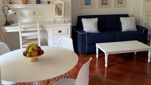 Un salon avec une table de salle à manger ronde, des chaises blanches, un canapé bleu orné de coussins à motifs, une table basse blanche et un ventilateur de plafond.