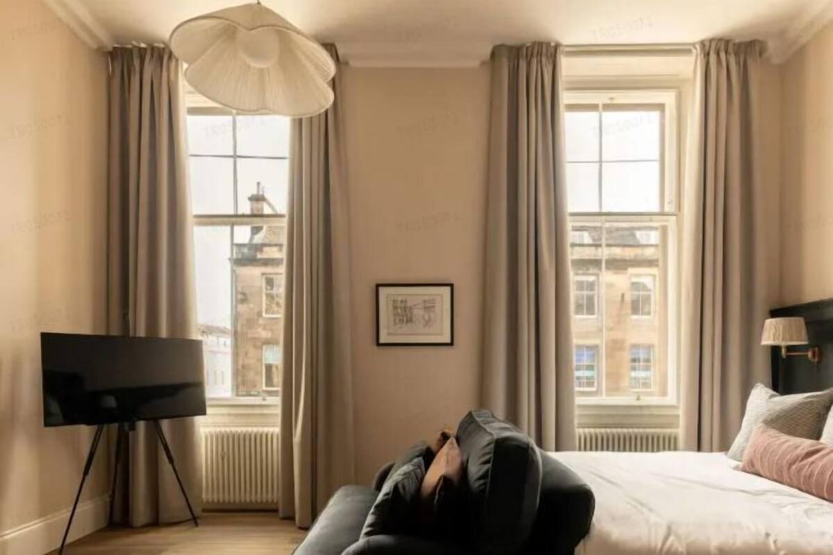 A hotel room with a bed, a TV on a stand, a window with curtains, and a radiator.