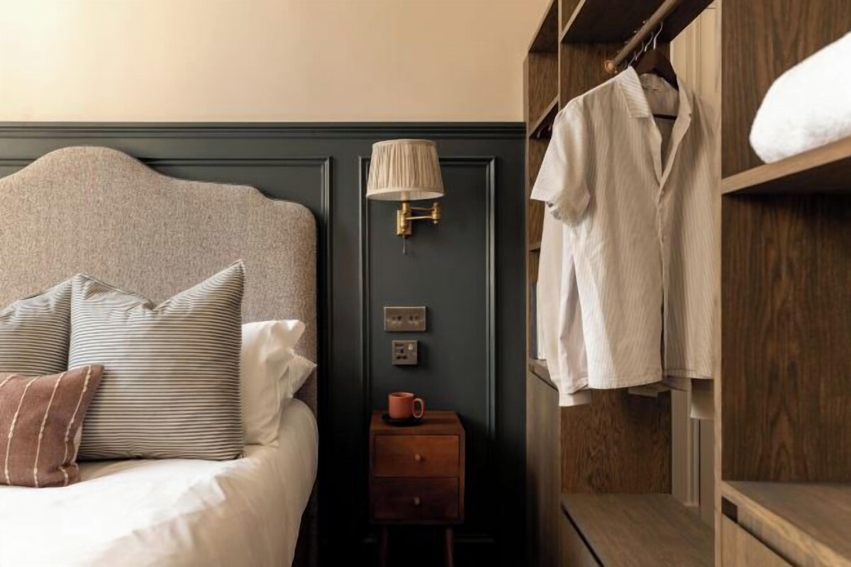 A neatly made bed with pillows, a bedside lamp, a small wooden nightstand with a cup, and a wardrobe with hanging clothes.