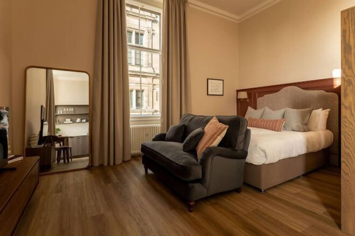 A hotel room with a bed, a sofa, a mirror, a window, and a wooden floor.