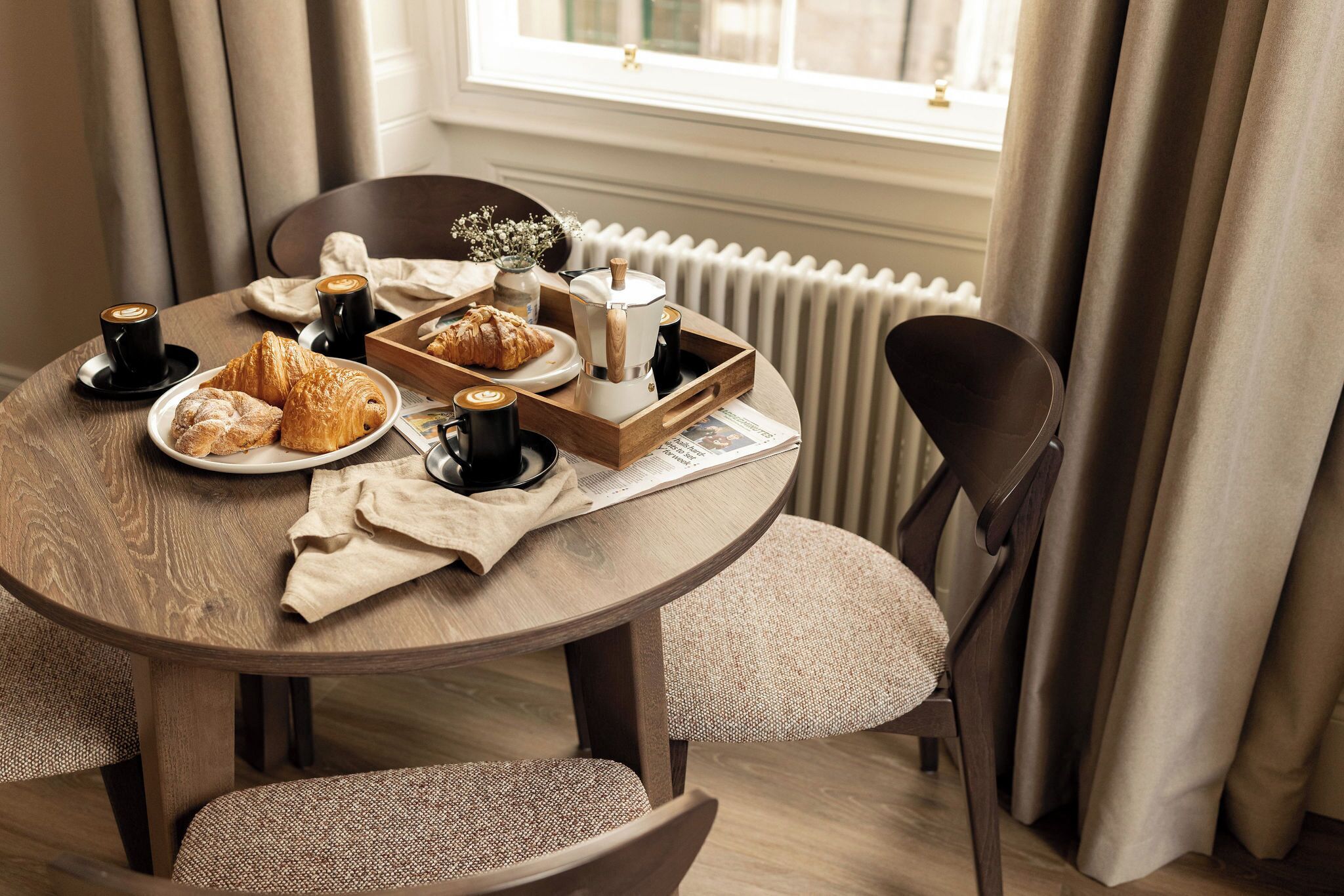 Une table de salle à manger ronde en bois, sur laquelle on trouve des assiettes de croissants et une tasse de café, une machine à café et un plateau rempli d’autres pâtisseries.