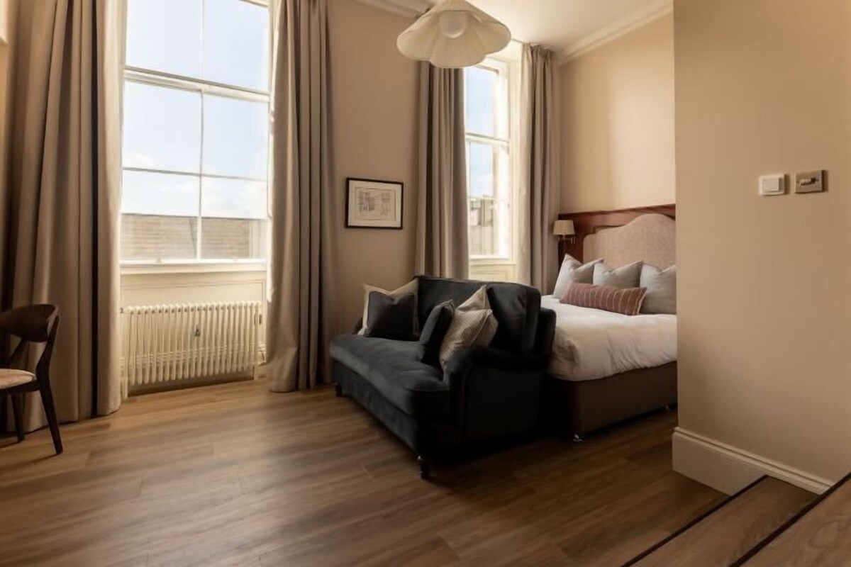 Una camera d'albergo con un letto, una poltrona, una sedia, un termosifone e una finestra con tende.
