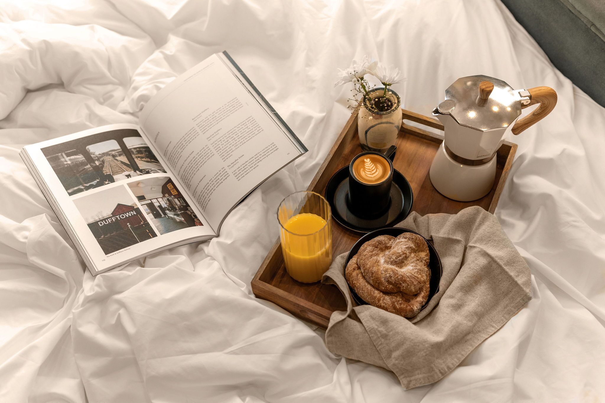 A breakfast setup on a bed with a book, a coffee pot, a glass of orange juice, and a plate of pastries.