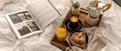 A breakfast setup on a bed with a book, a coffee pot, a glass of orange juice, and a plate of pastries.
