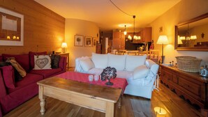 A cozy living room with a red sofa, a white sofa, and a wooden coffee table. There is a kitchen in the background with wooden cabinets and a hanging light fixture. - The flat is located above the village of Gryon, in Les Verneys Soleil, 2 km from the Fracherets ski (Gryon)