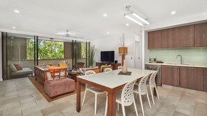 Eine moderne Küche mit Holzschränken, ein Essbereich mit weißem Tisch und Stühlen sowie ein Wohnbereich mit Sofa und Couchtisch.