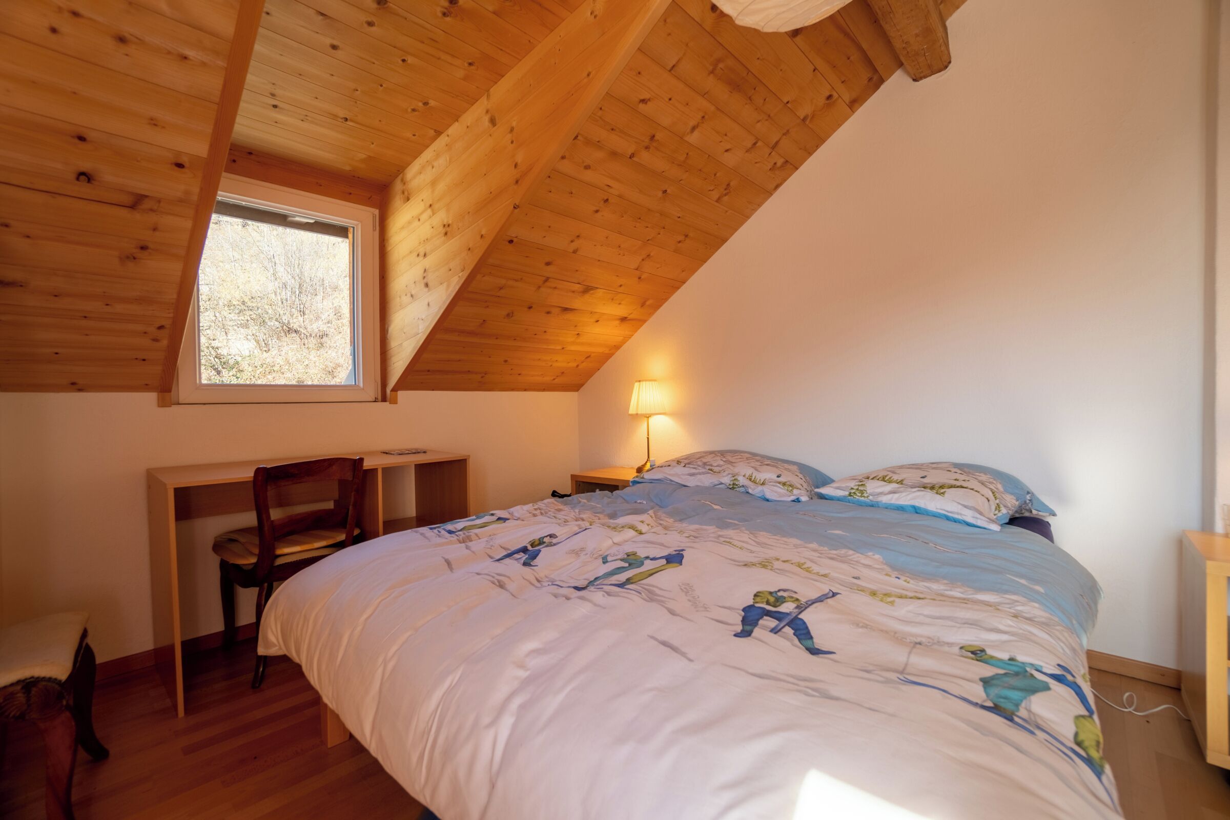 Une chambre avec un plafond en bois incliné, un bureau, une chaise, un lit avec une couette à motifs et une lampe de chevet.