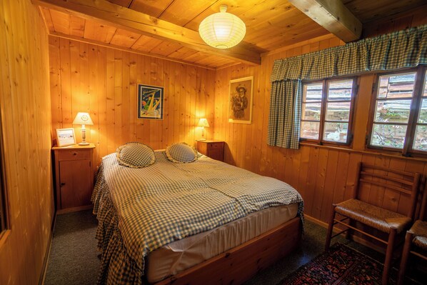 A wooden room with a bed, two bedside tables, a chair, and a window with curtains.