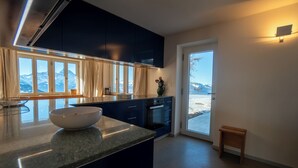 A modern kitchen with dark cabinets, a granite countertop, and a view of mountains through large windows. - The chalet, which has just been renovated in 2024, is located 200m from the ski slope linking the Vi (Gryon)
