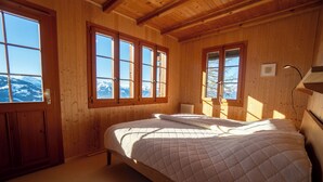 A wooden cabin bedroom with a bed, large windows, and a view of snow-capped mountains. - The chalet, which has just been renovated in 2024, is located 200m from the ski slope linking the Vi (Gryon)