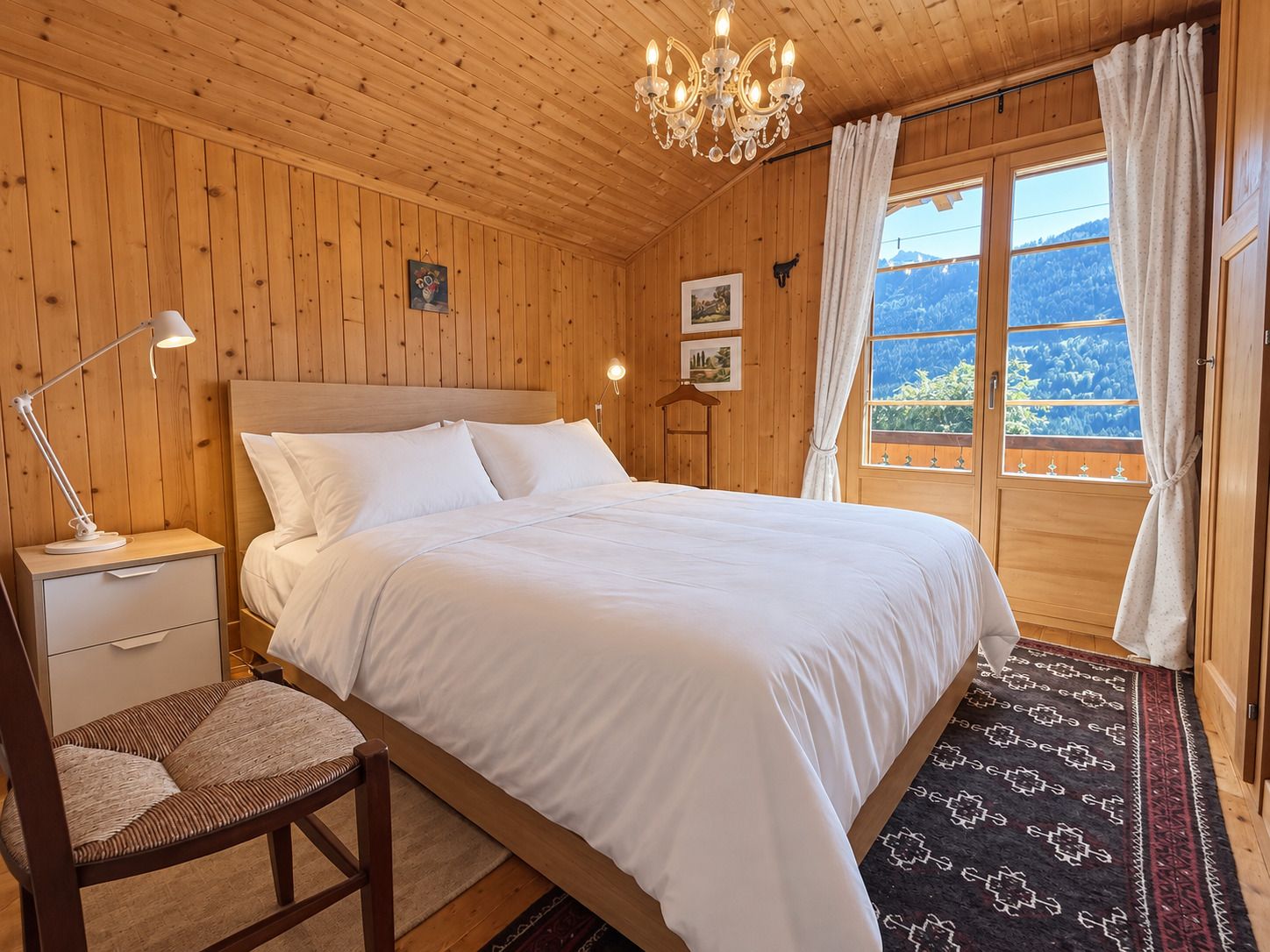 A wooden room with a bed, a nightstand, a chair, a chandelier, and a window with curtains.