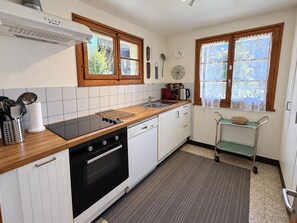 Una cocina con estufa negra, armarios blancos, encimera de madera, un carrito pequeño y una ventana con cortina estampada.