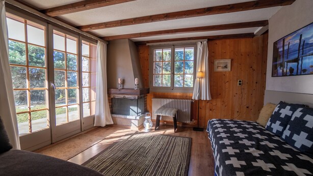 A cozy room with wooden beams, a large window with curtains, a bed with a patterned blanket, and a small table with a lamp.