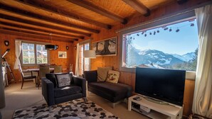 Una sala de estar acogedora con paredes y techo de madera, un ventanal con vistas a la montaña, un televisor de pantalla plana y un comedor con mesa y sillas.