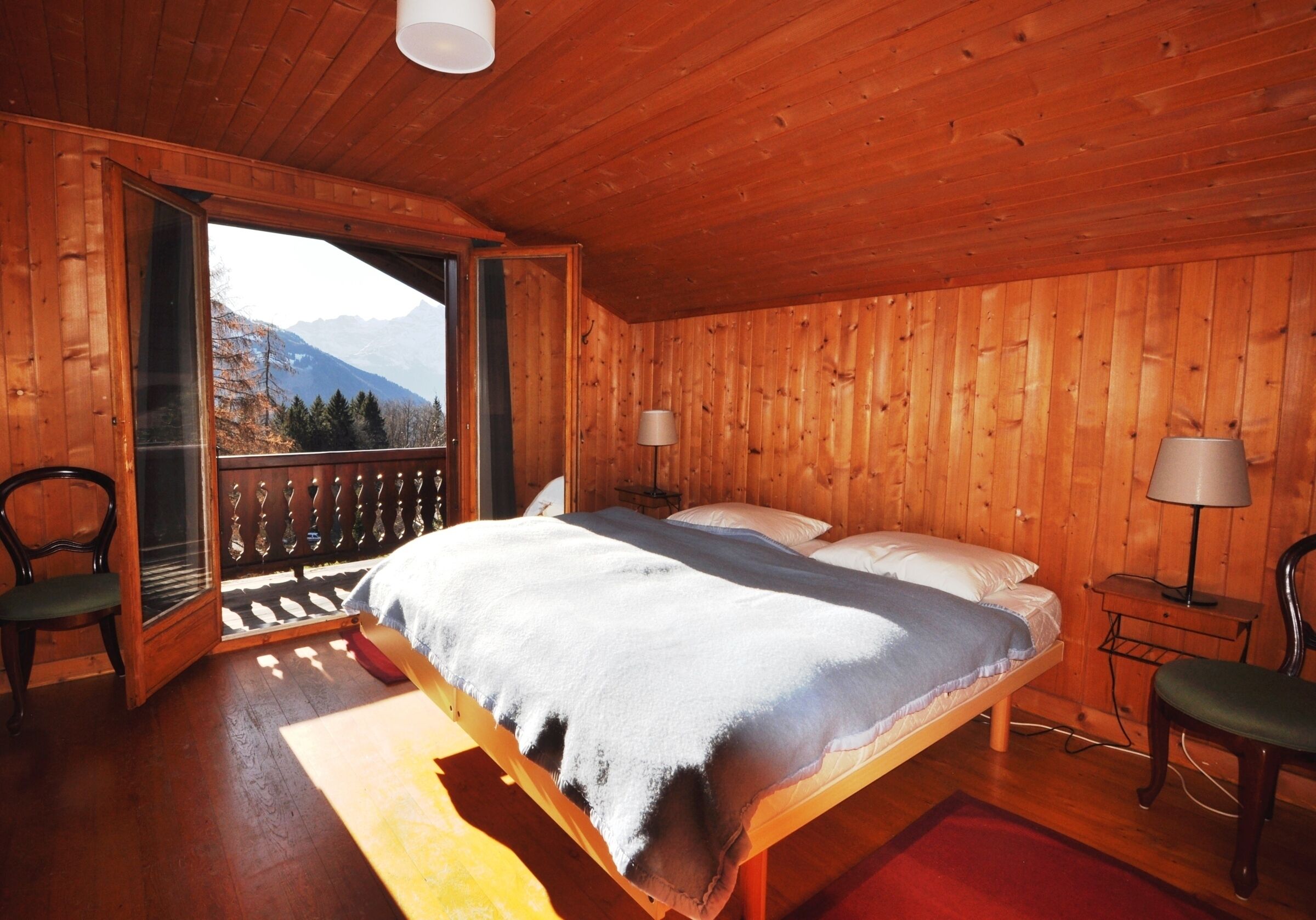 Ein Schlafzimmer einer Holzhütte mit einem Bett, zwei Stühlen, einem Nachttisch und einem Fenster mit Blick auf einen Berg.