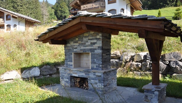 Una chimenea de piedra al aire libre con techo de madera, rodeada por una base de piedra y ubicada en una zona ajardinada con vista a una casa al fondo.