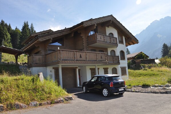 Un chalet de dos pisos con un balcón, un coche estacionado adelante y montañas al fondo.