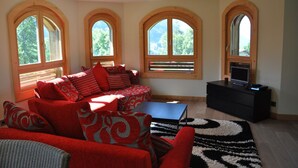 Una sala de estar con un sofá rojo, una mesa de centro negra y un mueble de entretenimiento negro. Hay tres ventanas arqueadas grandes que proporcionan luz natural y vistas a árboles.
