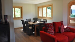 Un comedor con mesa y sillas de madera, un sofá rojo con cojines estampados y dos ventanas con marcos de madera.