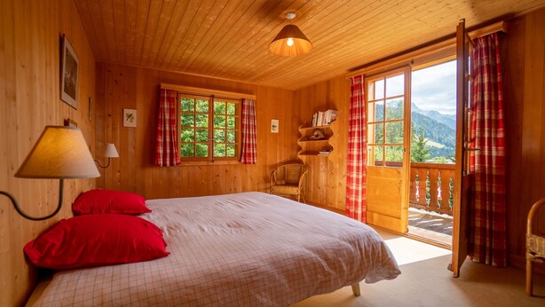 Un dormitorio de cabaña de madera con una cama, almohadas rojas, una lámpara, una silla y vistas a las montañas a través de un amplio ventanal.