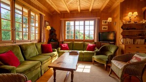 Una sala de estar acogedora con techo de madera, sofás verdes con cojines rojos, una mesa de centro de madera, sillas de mimbre y una estantería con lámparas.