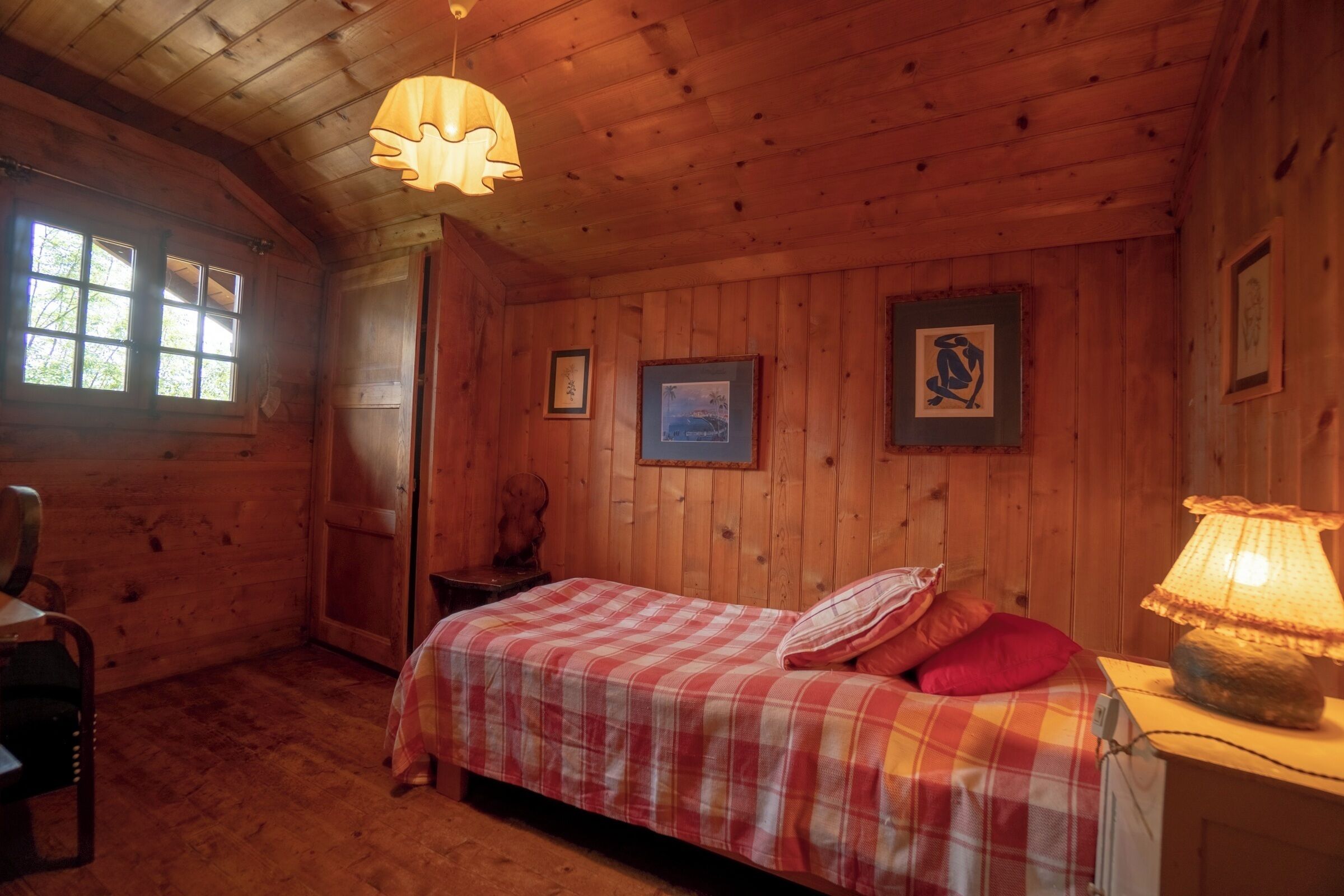 A cozy cabin room with a plaid bed, wooden walls, a hanging lamp, and framed pictures on the wall.
