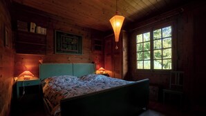 A cozy wooden cabin bedroom with a bed, two bedside lamps, a window with a view of trees, and a hanging light fixture.