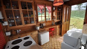 A rustic kitchen with wooden cabinets, a stove, a refrigerator, and a window with a red checkered curtain.