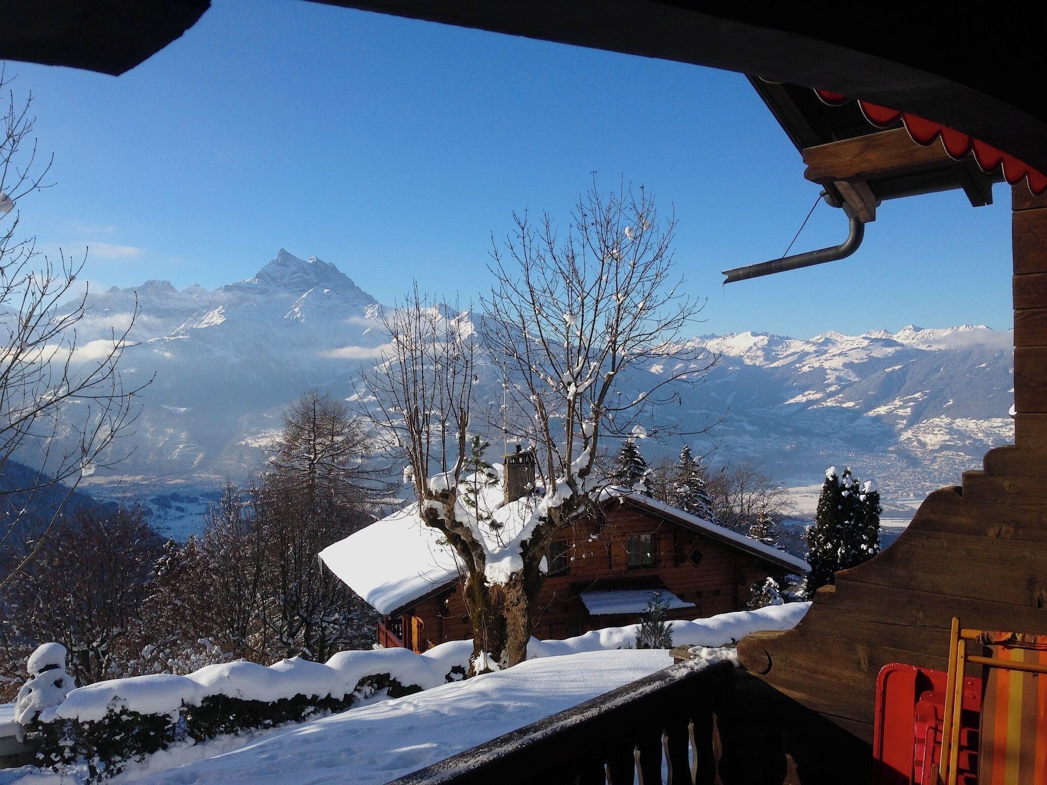 A wooden cabin with a snow-covered roof, surrounded by snow-covered trees and mountains in the background.