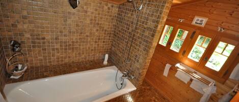 A bathroom with a large bathtub, tiled walls, wooden floors, and a window with a view of greenery.