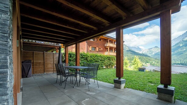 Un patio al aire libre con mesa y sillas, un área cubierta con vigas de madera, y vistas a montañas y un lago.