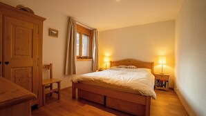 Un dormitorio con cama de madera, dos lámparas de noche, una ventana con cortinas, un armario de madera y una pequeña silla de madera.