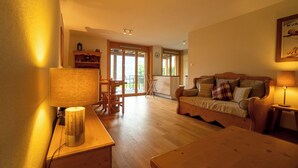 Una sala de estar acogedora con un sofá de madera, una mesa de comedor y una lámpara. Se ve un área de cocina a través de una puerta abierta.