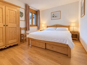 A bedroom with a wooden bed, a wooden wardrobe, a chair, a bedside table with a lamp, and a window with curtains. - The apartment is located 150m from the Place de la Barboleuse in Gryon and is close to all amenities (Gryon)