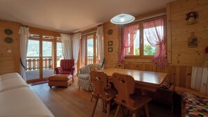 Interior de una cabaña de madera con sofá, una mesa de comedor con sillas y una ventana con cortinas.