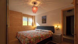 Un dormitorio con suelo de madera, una cama con colcha estampada, una mesita de noche con lámpara y una ventana con cortinas.