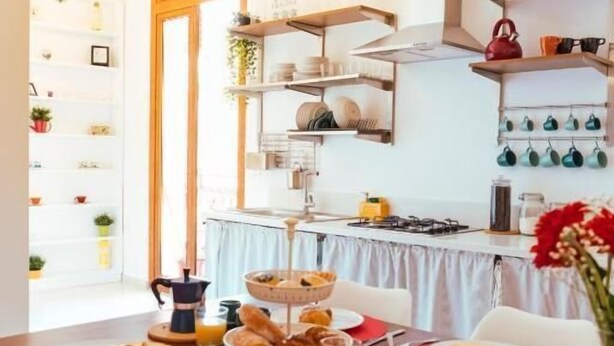 A dining area with a wooden table set for breakfast, a kitchen in the background, and shelves with kitchenware. - Relais CuoreNapoletano (Naples)