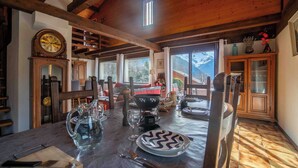 Un comedor con una mesa de madera dispuesta para dos personas, un reloj grande en la pared y vistas a montañas a través de las ventanas.