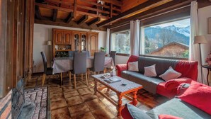 Una sala de estar acogedora con techo de madera, un sofá con cojines rojos, un comedor con mesa y sillas, y vistas a las montañas a través de amplios ventanales.