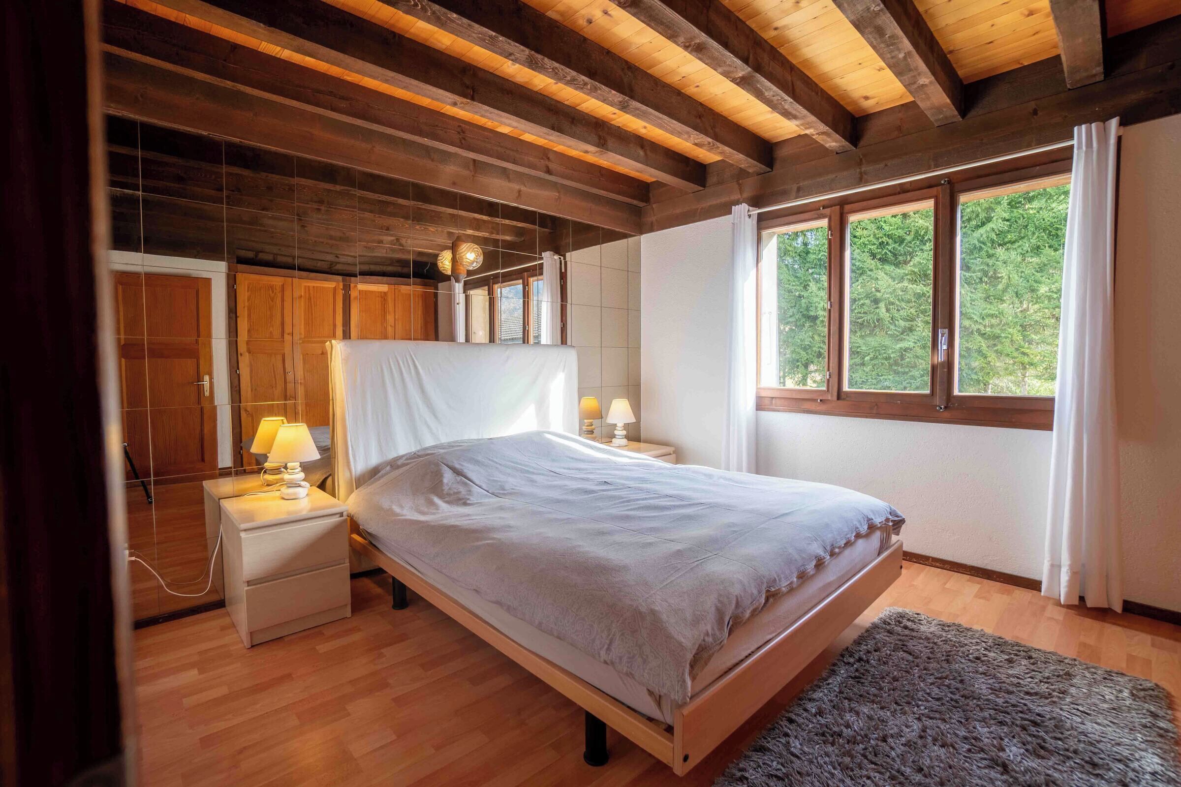 A bedroom with a wooden bed, nightstands with lamps, a large window with curtains, and a wooden ceiling.
