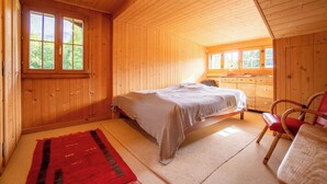 Dormitorio de cabaña de madera con una cama, una silla, un aparador y una ventana con vistas a árboles.