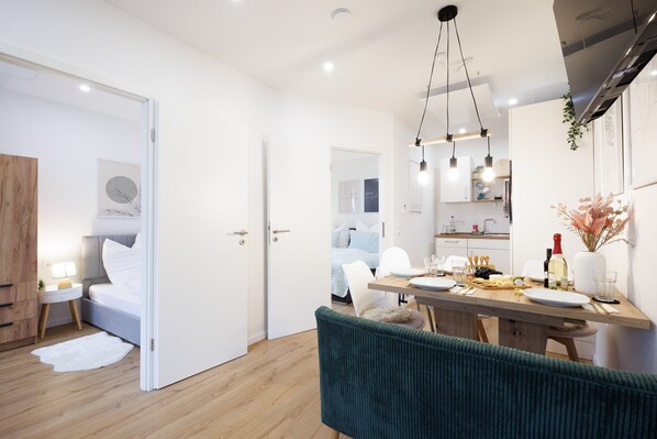Una habitación moderna y luminosa con una mesa de comedor de madera, un banco tapizado de verde y una cama blanca con cabecera de madera.