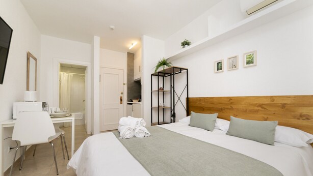 A hotel room with a bed, a desk with a chair, a mirror, and a shelf with plants. - Classy IX - Ferga Suites (Madrid)