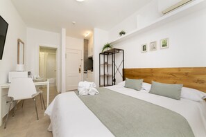 A hotel room with a bed, a desk with a chair, a mirror, and a shelf with plants. - Classy IX - Ferga Suites (Madrid)