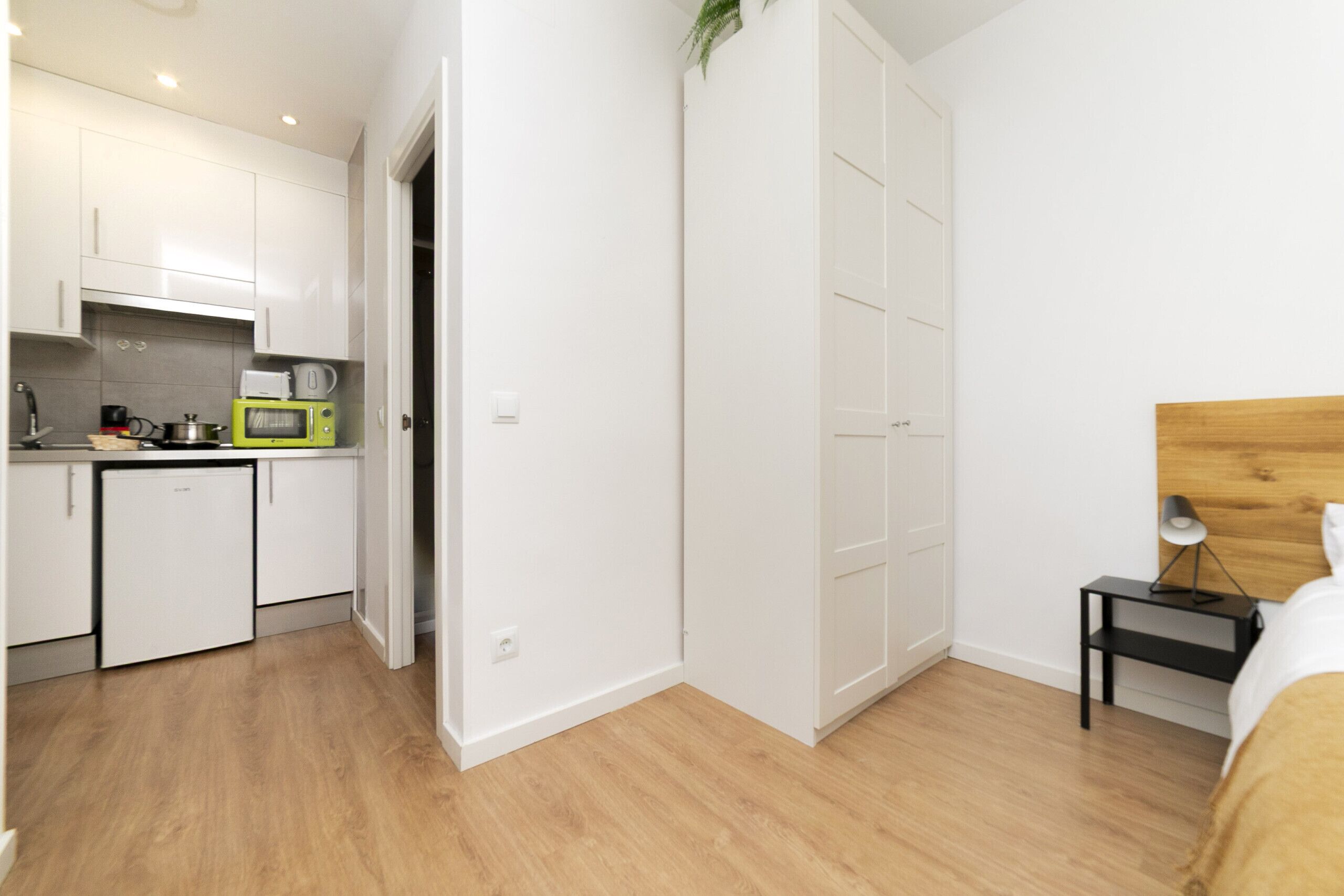 A compact kitchen with white cabinets, a microwave, and a sink. There is a wooden floor and a bed with a wooden headboard in the adjacent room.