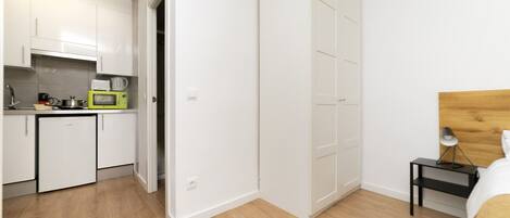 A compact kitchen with white cabinets, a microwave, and a sink. There is a wooden floor and a bed with a wooden headboard in the adjacent room.