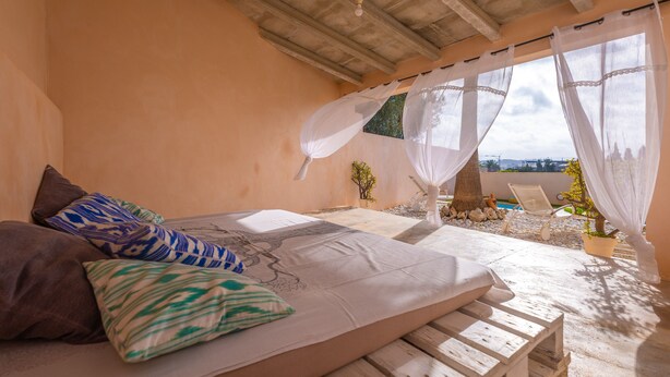 A bedroom with a bed, pillows, a wooden bench, a small table, and a view of the sea through sheer curtains.