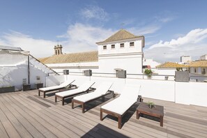 A rooftop terrace with sun loungers and a table, overlooking a cityscape with a prominent tower.