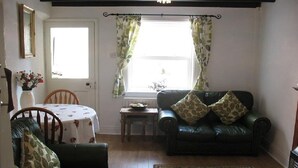 Una sala de estar acogedora con un sofá de cuero verde, una silla de madera, una mesita con mantel de flores y una ventana con cortinas verdes.