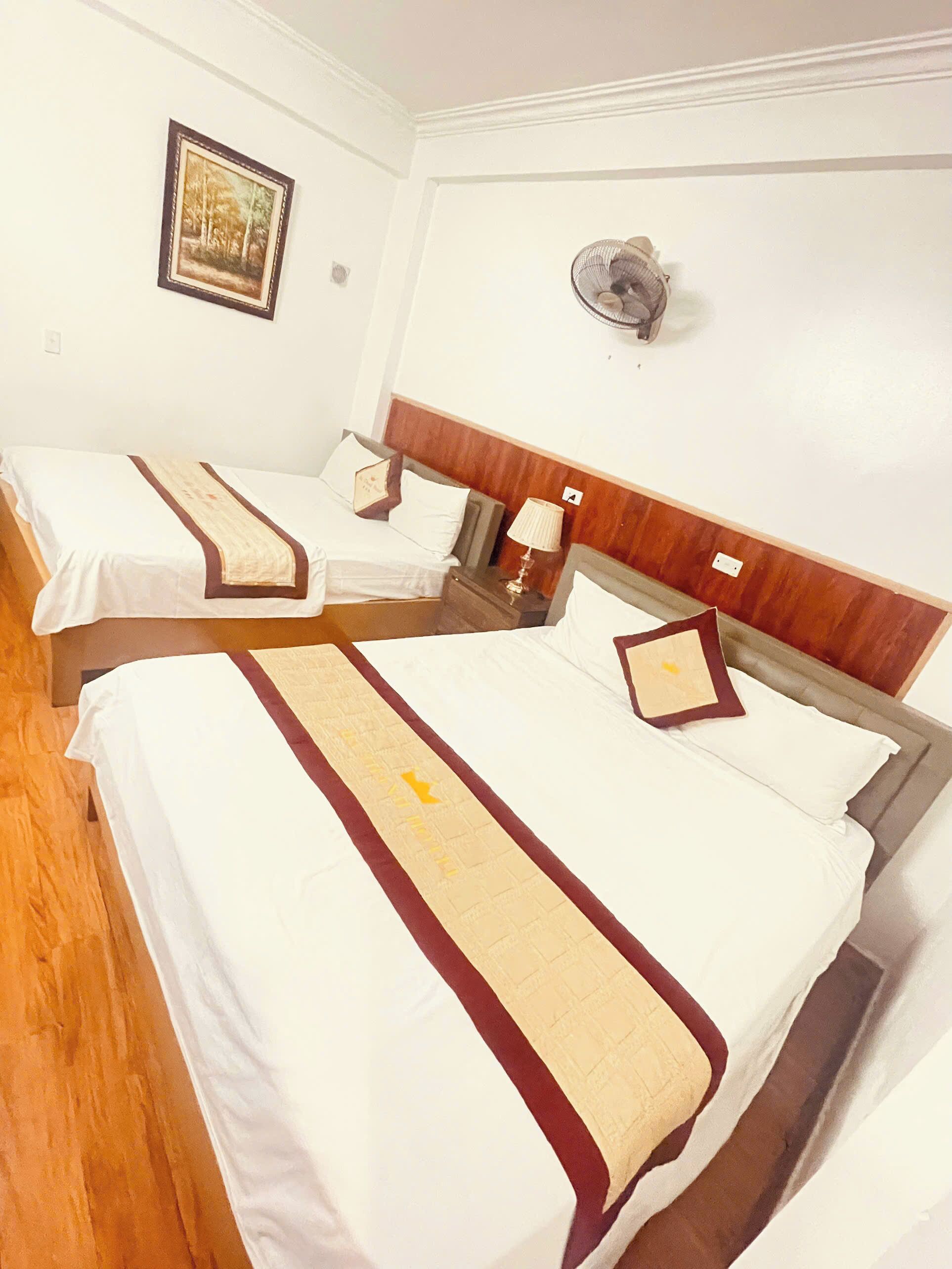 A hotel room with two beds, each with a wooden headboard, white bedding, and a decorative red and beige runner. There is a wall-mounted fan and a framed picture on the wall.